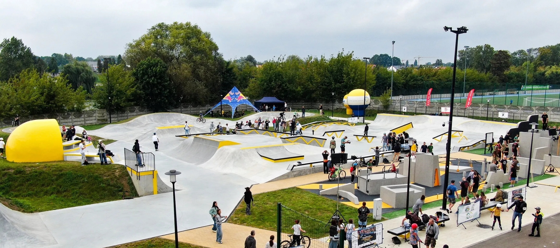 Skatepark z betonowymi rampami i przeszkodami, na którym obecnych jest wiele osób jeżdżących na rowerach, hulajnogach i deskorolkach oraz widzowie.